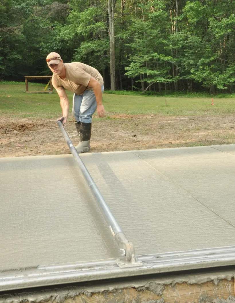 Archival photograph: a worker walks a bull float across a fresh slab poured up against a mown lawn on a bright day.
