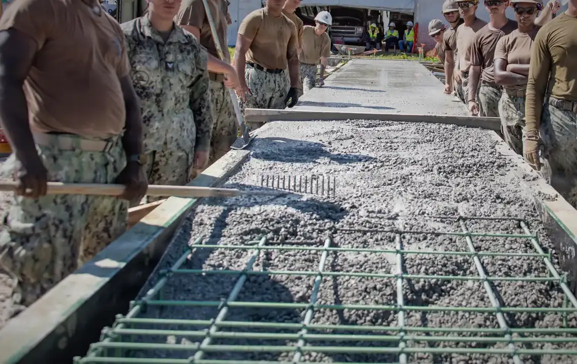 Archival photograph: a mat of tied rebar sitting on chairs above a packed gravel base inside set wooden forms, before any concrete has been placed.