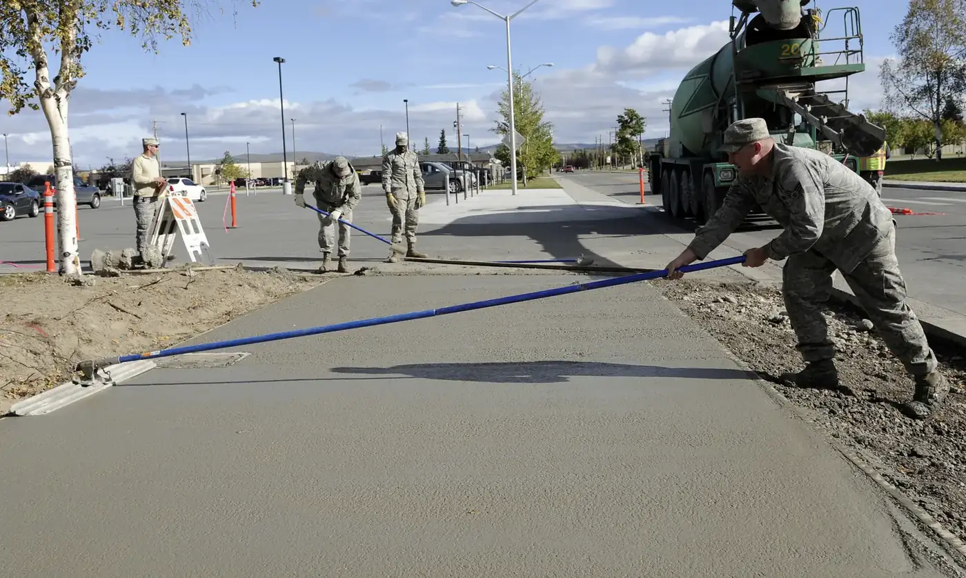 Archival photograph: a crew pulls a screed across a wide fresh slab beside a street, a mixer truck parked at the far end and the gravel shoulder still open along the edge.